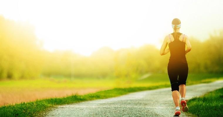 Na zdjęciu widać plecy biegnącej kobiety. Na zieloną okolicę pada światło wschodzącego słońca