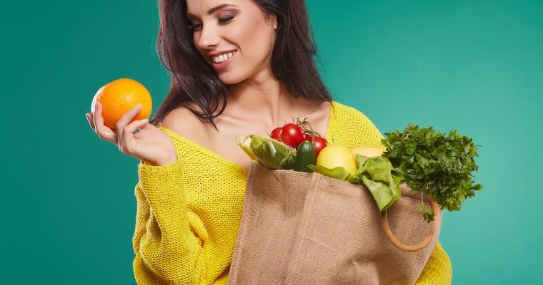 Młoda kobieta trzyma w lewej ręce torbę z warzywami a w prawej pomarańcz