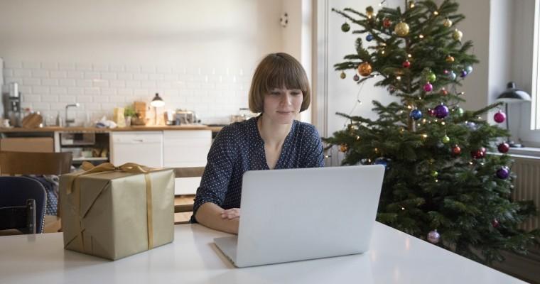 Kobieta siedzi przy stole przed laptopem. W tle choinka - kobieta kupuje prezenty przez internet. 