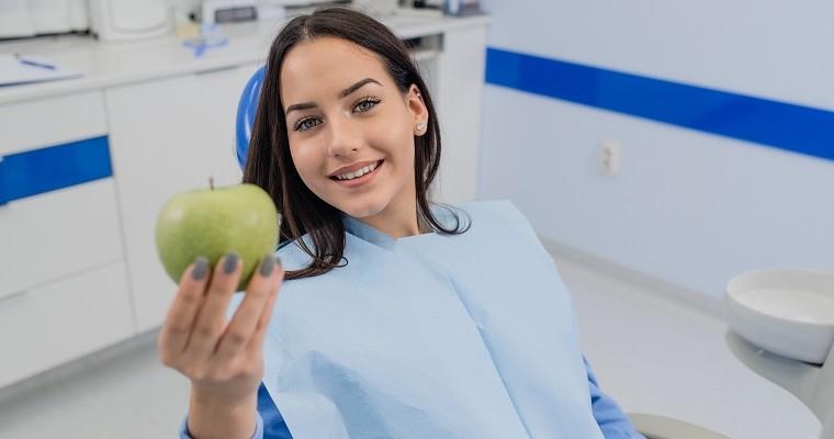 Kobieta siedzi na fotelu u stomatologa i trzyma w dłoni zielone jabłko 