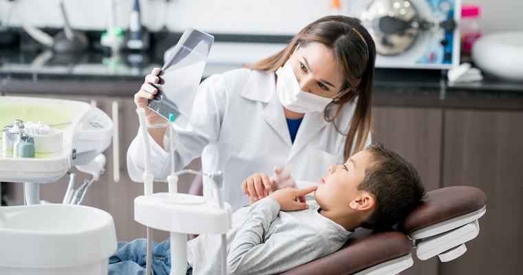 uśmiechnięta pani stomatolog ogląda zdjęcie rentgenowskie z małym chłopczykiem lezącym na fotelu w gabinecie  