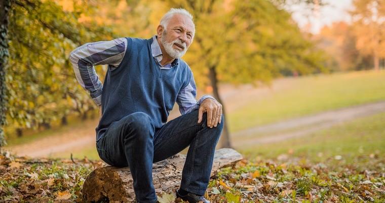 Starszy mężczyzna siedzi na pniu drzewa i trzyma się za bolące plecy