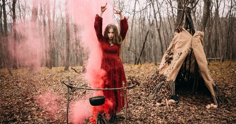 Młoda kobieta w czerwonej sukni stoi w lesie. Wzwnosi ręce do góry w tajemniczym geście. Obok znajduje się ognisko z którego bucha czerwona para