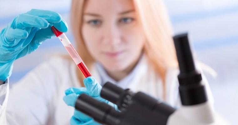 Kobieta w rękawiczkach i z mikroskopem przygląda się fiolce z krwią